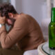 A desperate man in brown clothes sits near a bottle of alcohol and leans his head on his hands