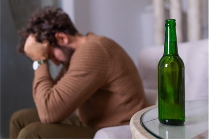 A desperate man in brown clothes sits near a bottle of alcohol and leans his head on his hands